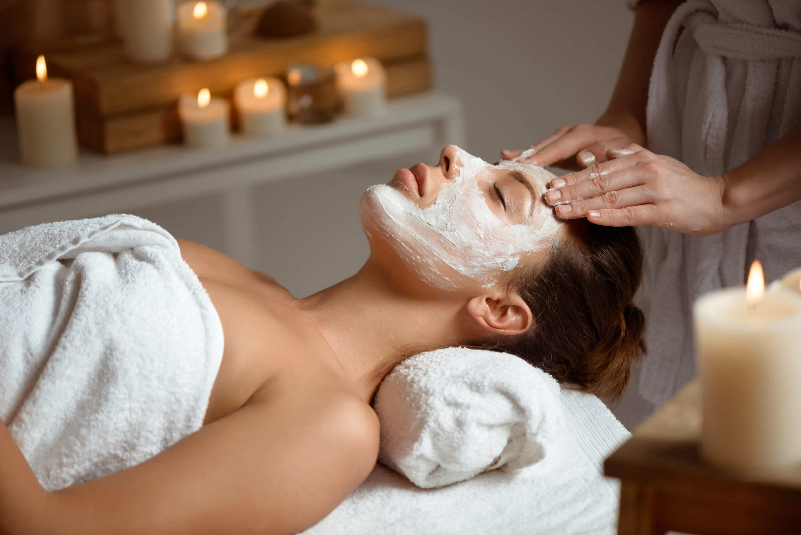 A woman relaxing during a professional facial treatment at a Florida med spa, with a white mask applied and soft candlelight creating a calming atmosphere.